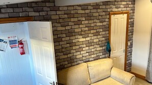 A room with a beige sofa, a brick-patterned wall, a white door, and a fire extinguisher on the wall. - Lovely 1-bed Chalet in Mablethorpe (Mablethorpe)