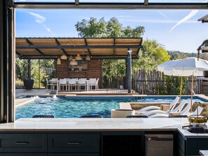A pool area with lounge chairs, a covered seating area, and a view of the surrounding greenery. - 10 Bedroom | Pool | Hot tub | Pickleball | Firepit (Dripping Springs)