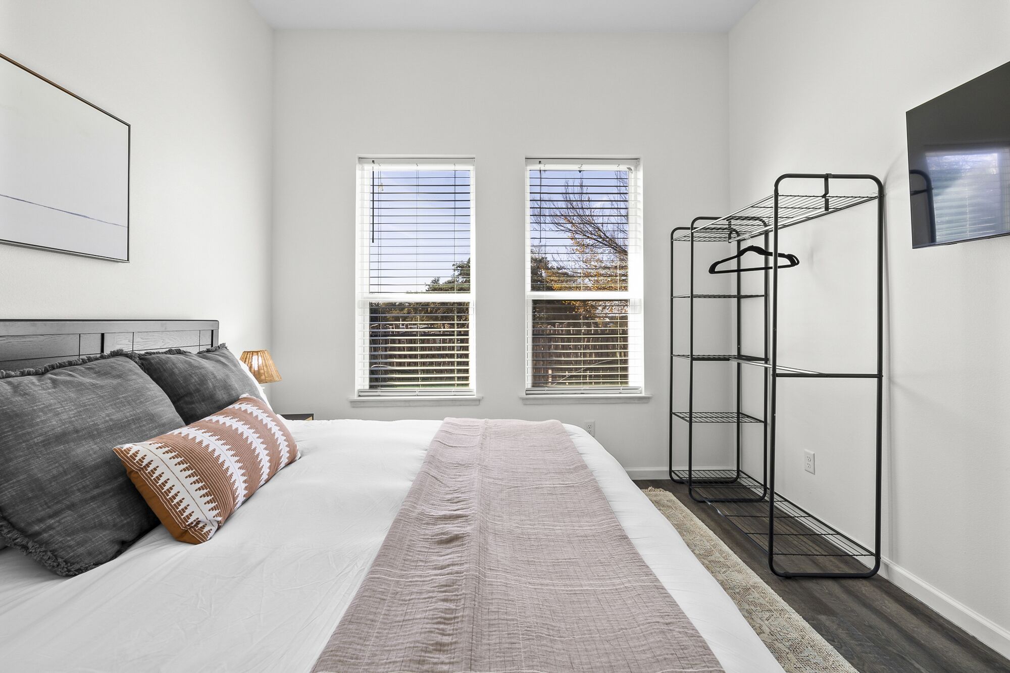 Un dormitorio con una cama, dos ventanas, un tendedero de metal y un cuadro en la pared.