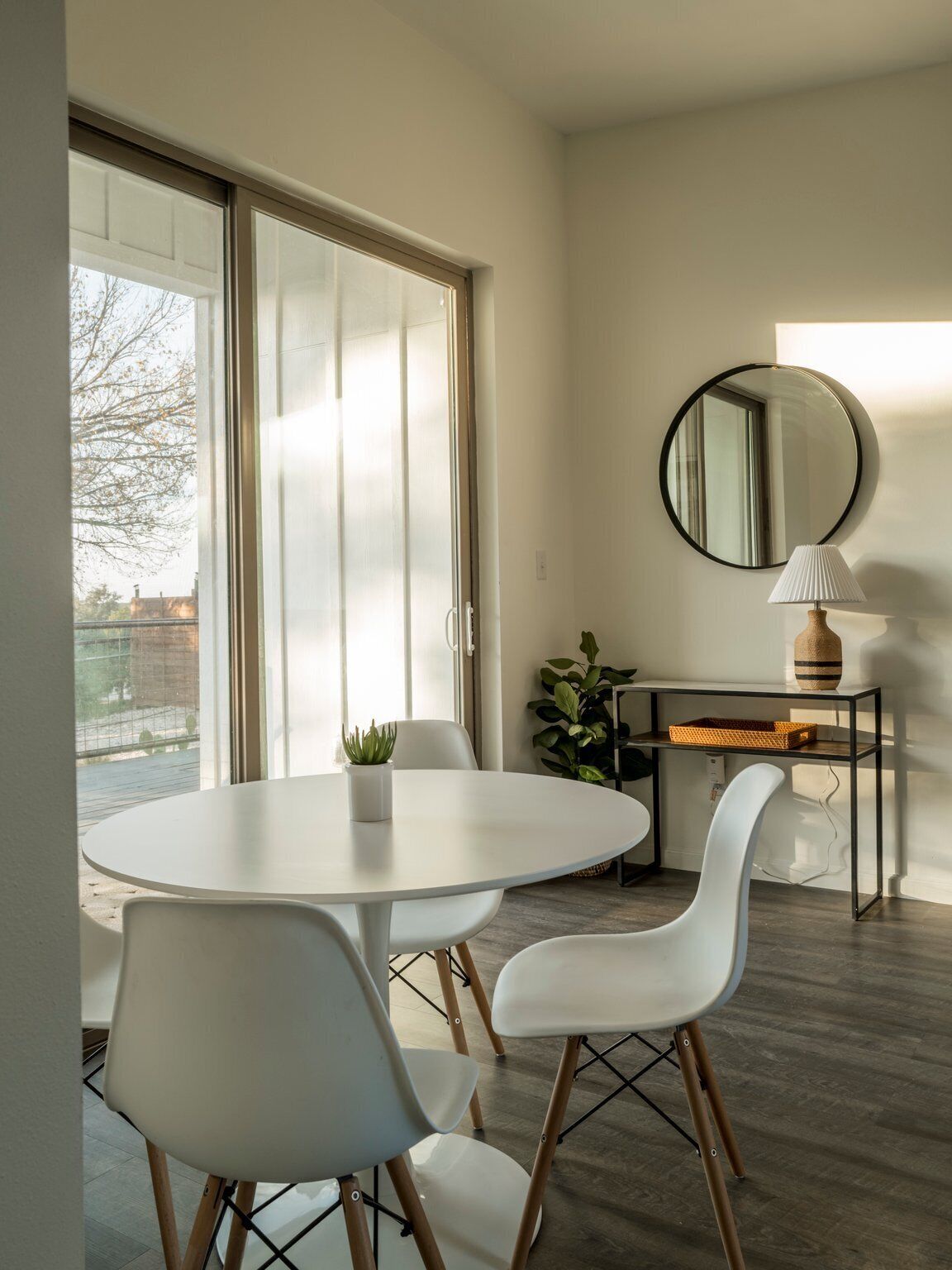 A modern dining area with a round table and white chairs, a desk with a lamp, and a large mirror on the wall.