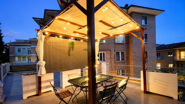A covered outdoor seating area with a table and chairs, a pergola with string lights, and a view of residential buildings in the background. - Lumina Hidden Terrace - Charming Family Apartment in the Heart of Perugia (Perugia)