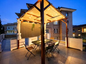 Un espace extérieur aménagé, abrité, avec une table et des chaises, une pergola décorée de guirlandes lumineuses, et une vue sur des immeubles d’habitation en arrière-plan.