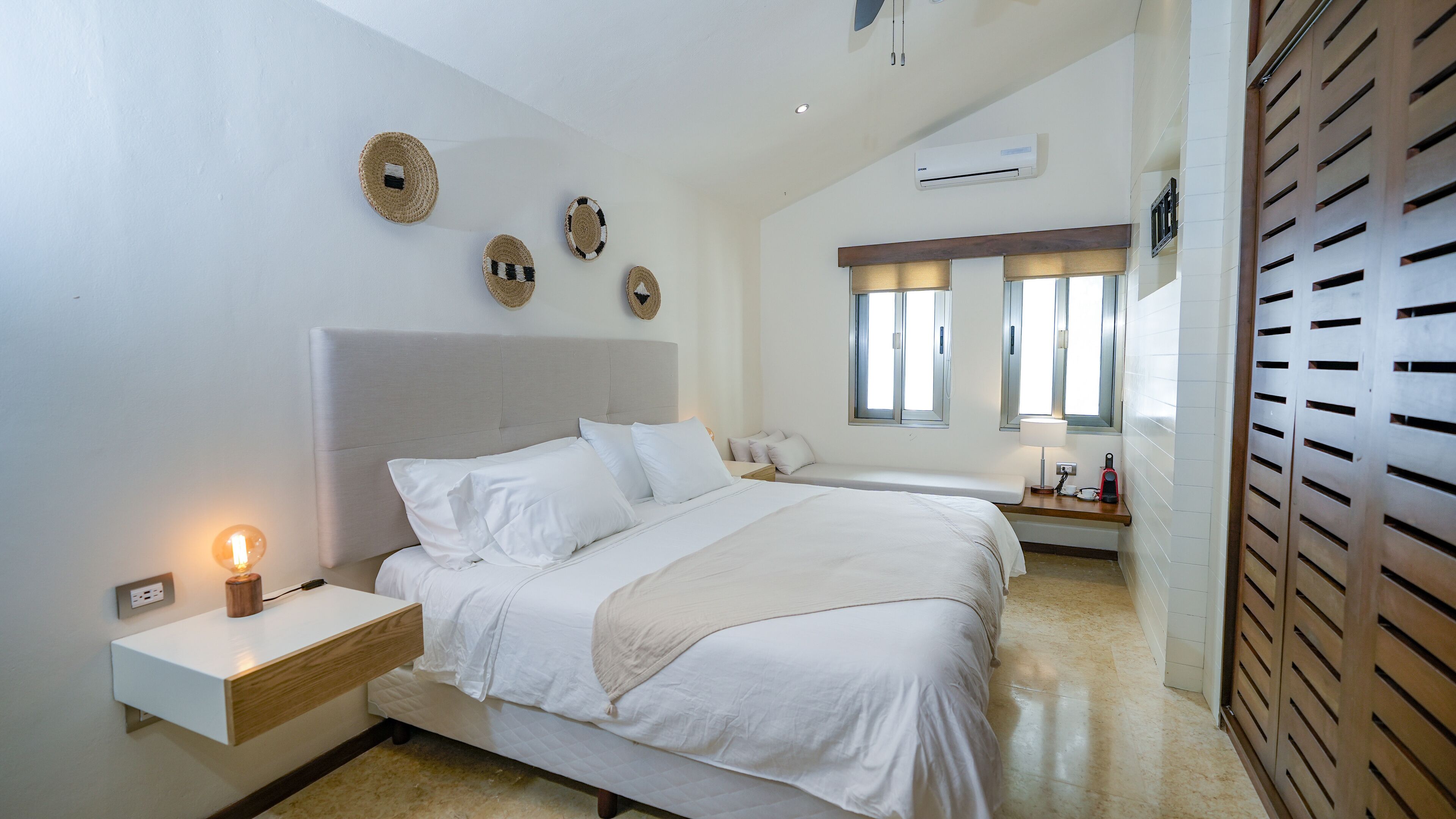 A modern bedroom with a large bed, bedside tables, a nightstand, a lamp, and a wall-mounted air conditioner.