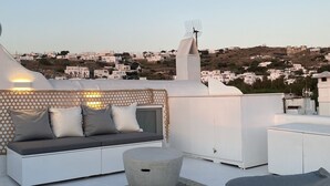 A rooftop terrace with a sofa set, a round table, and a view of a hillside town. - Villa Loom Mykonos Town (Mykonos)