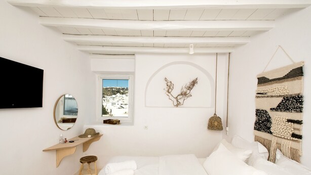A minimalist bedroom with a bed, a bedside table, a mirror, and a window with a view of buildings. - Villa Loom Mykonos Town (Mykonos)