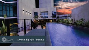 A modern swimming pool area with a sunset view, surrounded by a minimalist building with large windows and outdoor seating.