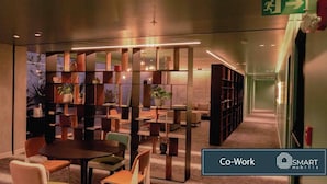 A modern co-working space with a central table and chairs, surrounded by bookshelves and potted plants.