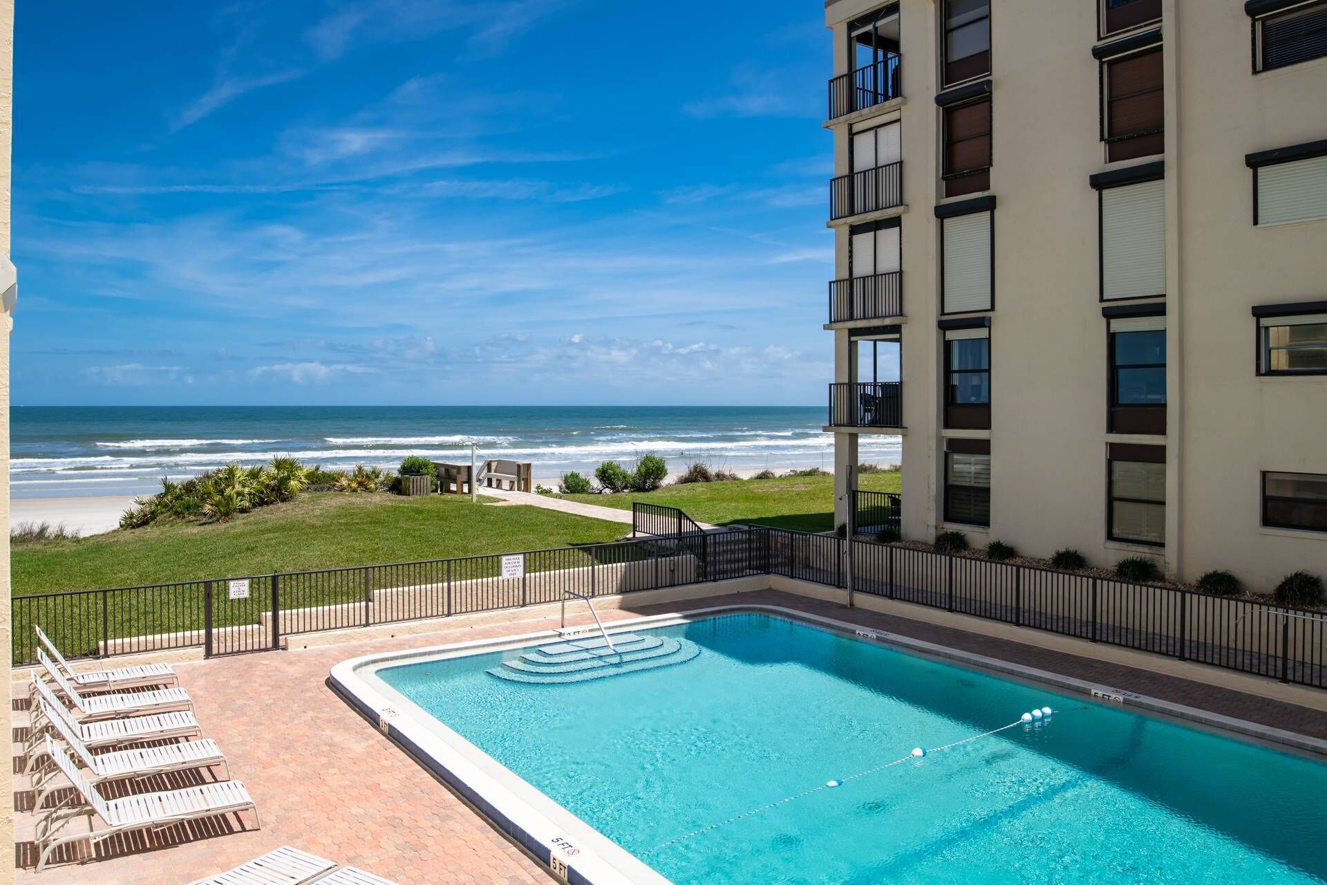 A swimming pool with a view of the ocean, a grassy area, and a building with multiple balconies.