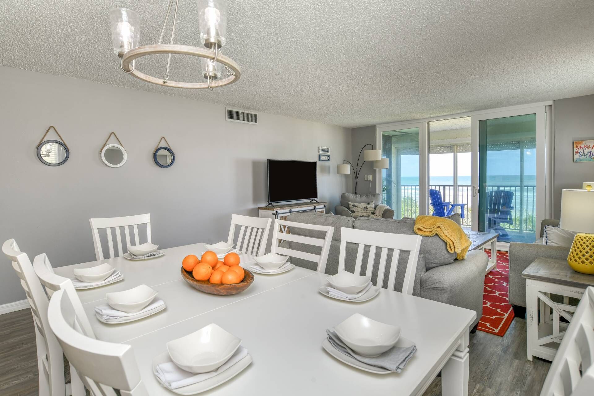 A dining area with a white table set for four, a bowl of oranges, and a TV in the background.