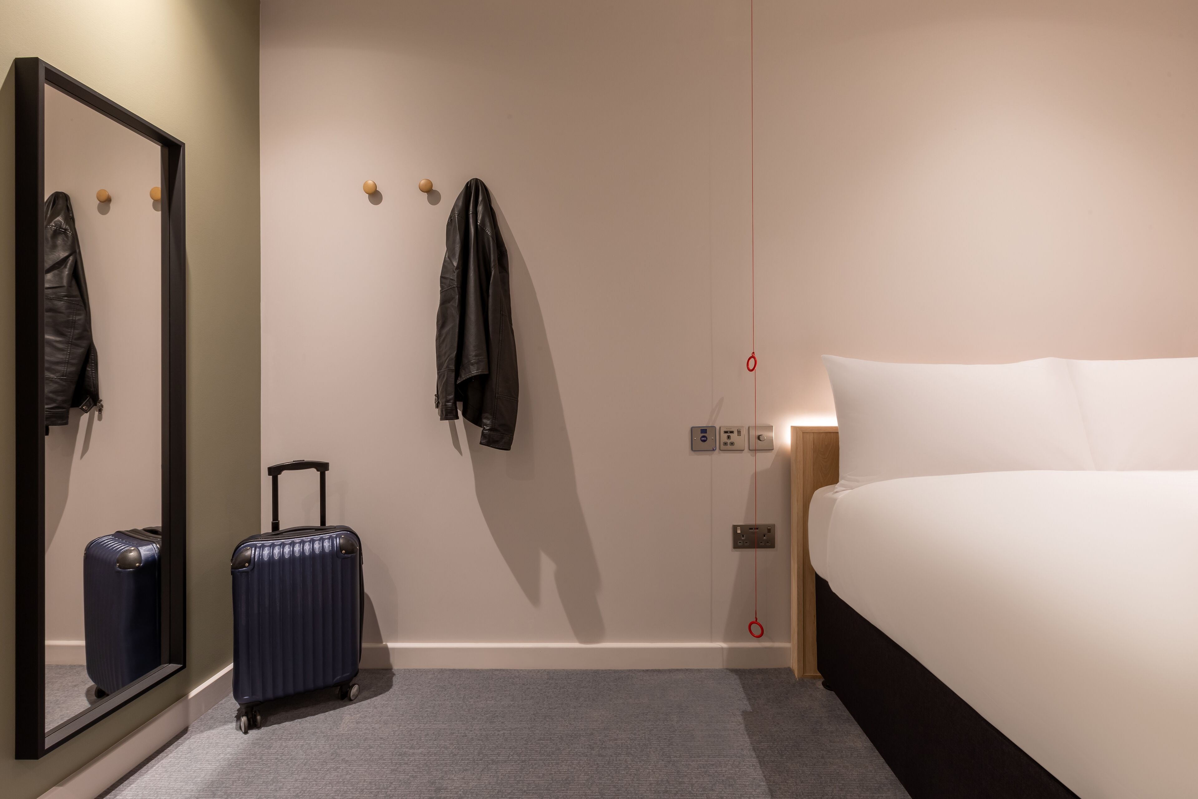 Habitació d'hotel amb un llit, una maleta, una jaqueta penjada a la paret i un mirall.