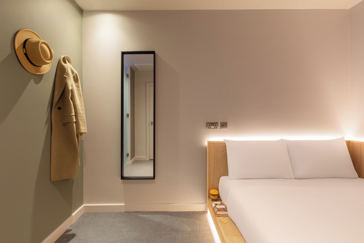 A hotel room with a bed, a nightstand, a mirror, a hat, and a coat hanging on the wall.