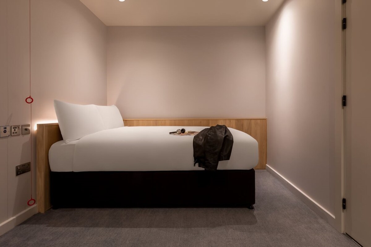 A modern hotel room with a single bed, a wooden headboard, and a grey carpet.