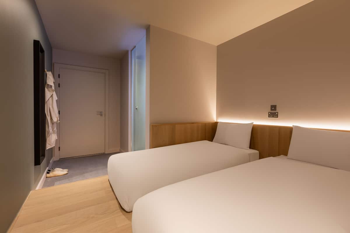 A hotel room with two beds, a wooden headboard, a white wall, and a door.