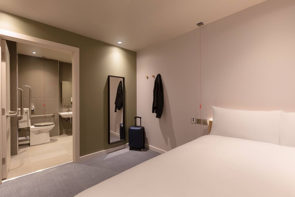 A hotel room with a bed, a suitcase, a mirror, and a bathroom visible through an open door.
