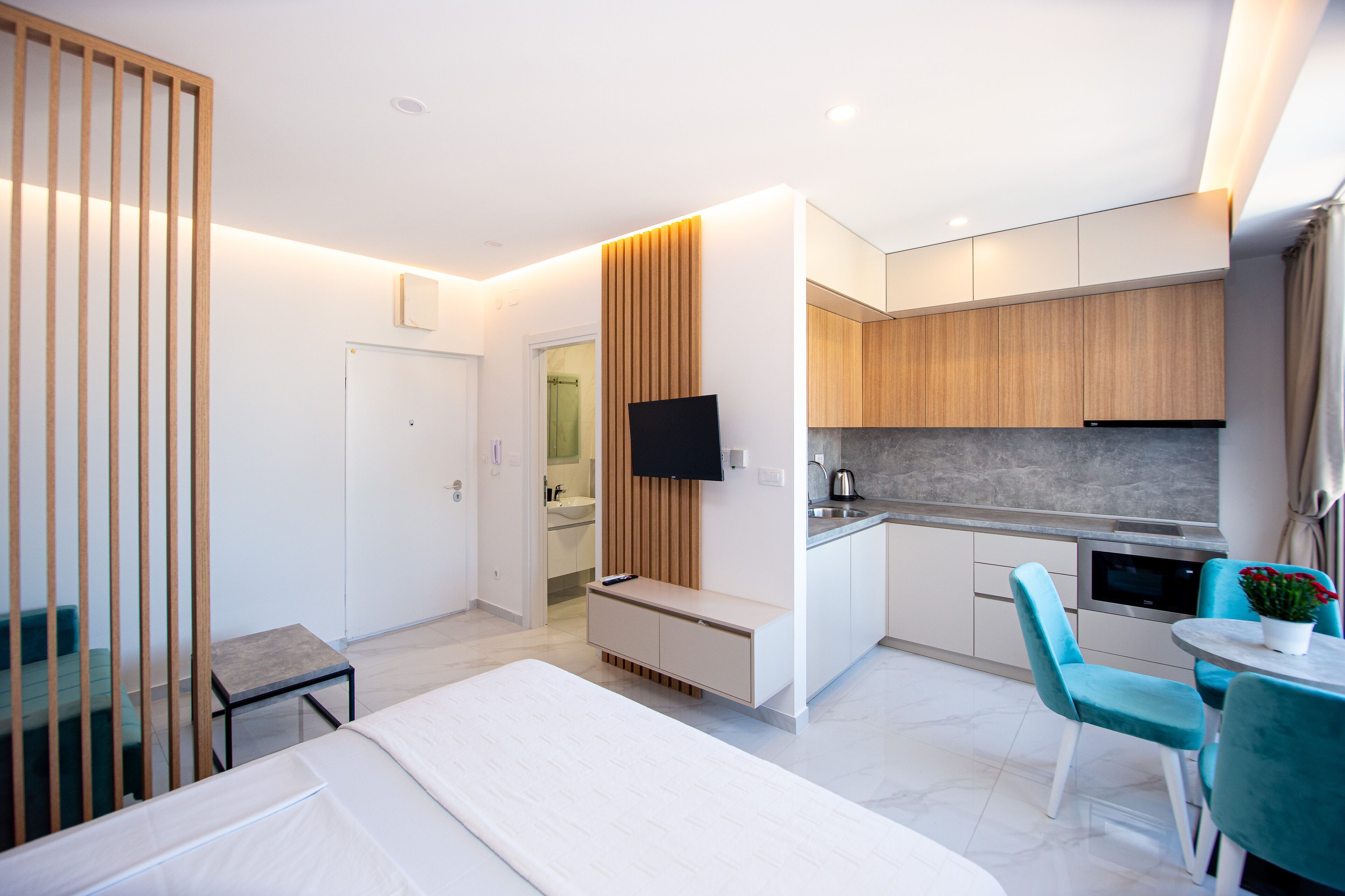 A modern hotel room with a bed, a small table, a chair, a TV, a kitchenette, and a bathroom.