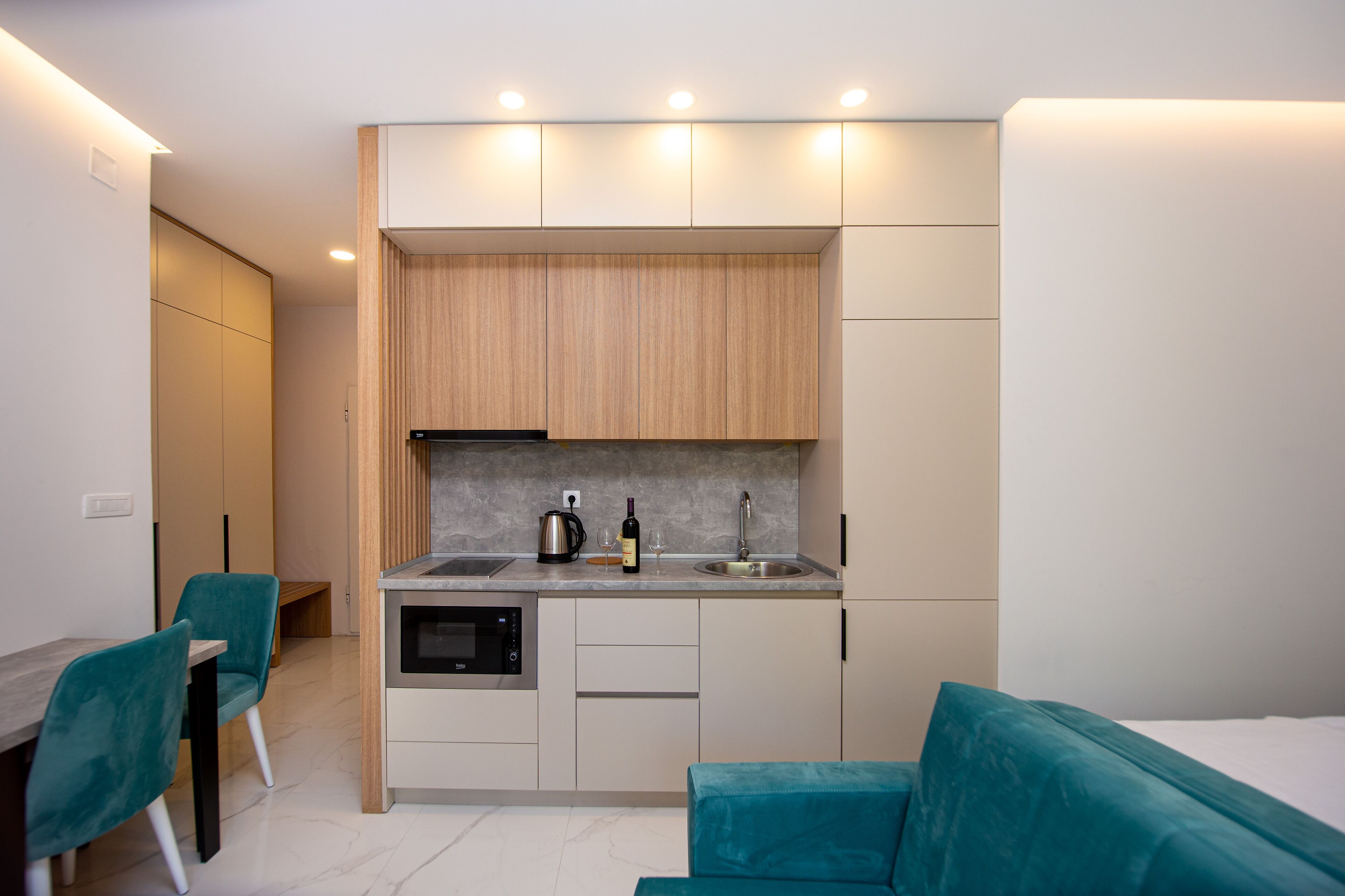 A compact kitchen with a built-in oven, a sink, and a countertop with a kettle and wine bottle. There are teal chairs and a teal sofa in the living area.