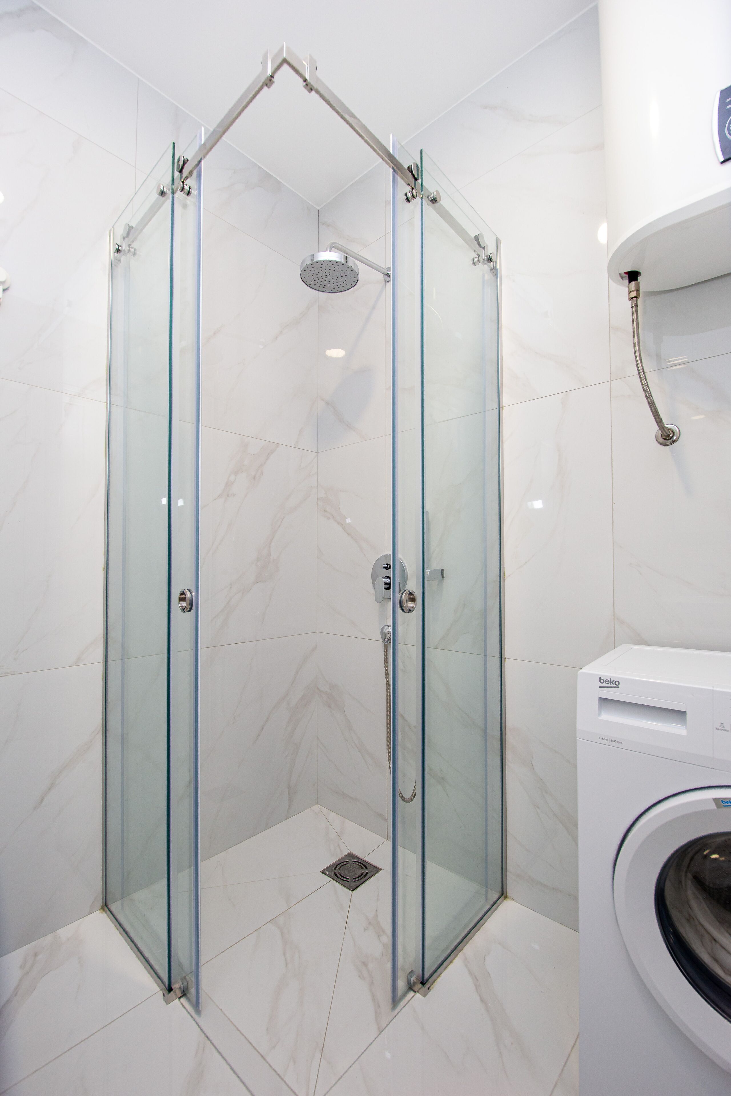 Una ducha con cerramiento de vidrio, un cabezal de ducha manual, un perchero en la pared y una lavadora en un baño con baldosas de mármol blanco.