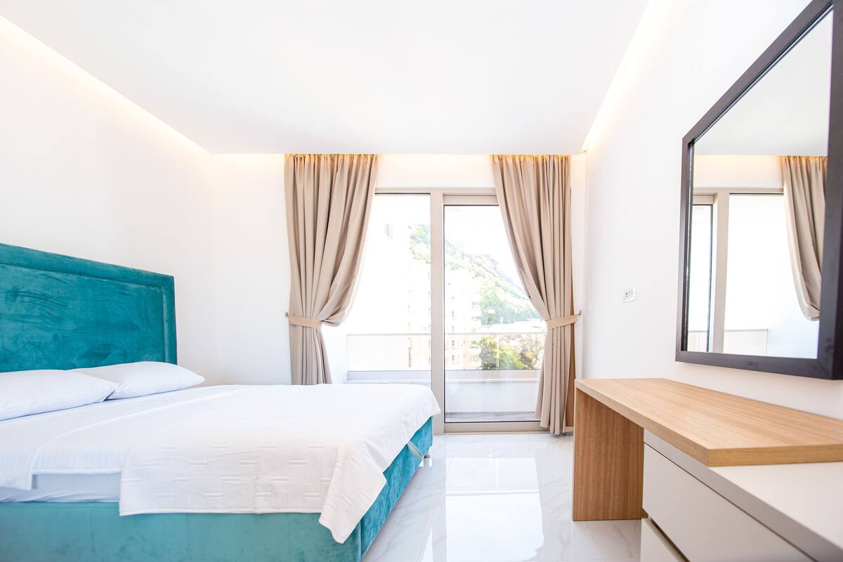 A modern bedroom with a teal-colored bed, white bedding, a wooden bedside table, and a large mirror.