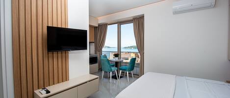 Une chambre d’hôtel avec un lit, un téléviseur fixé au mur, une petite table avec des chaises et une vue sur la mer grâce à de grandes fenêtres.