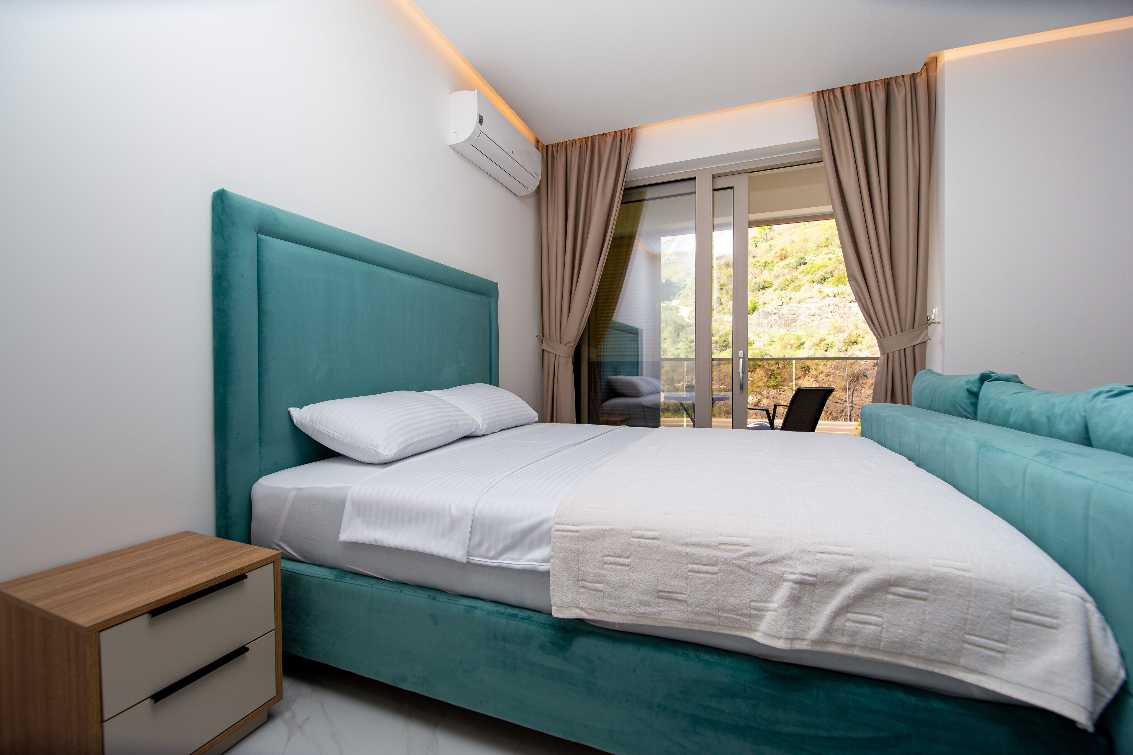 A modern bedroom with a teal bed, white linens, a wooden nightstand, and a view of the outdoors through a sliding glass door.