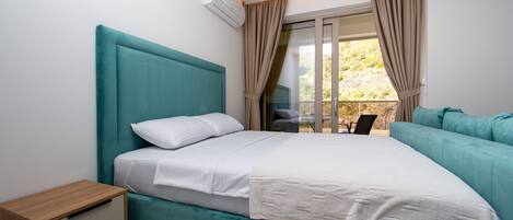 A modern bedroom with a teal bed, white linens, a wooden nightstand, and a view of the outdoors through a sliding glass door.