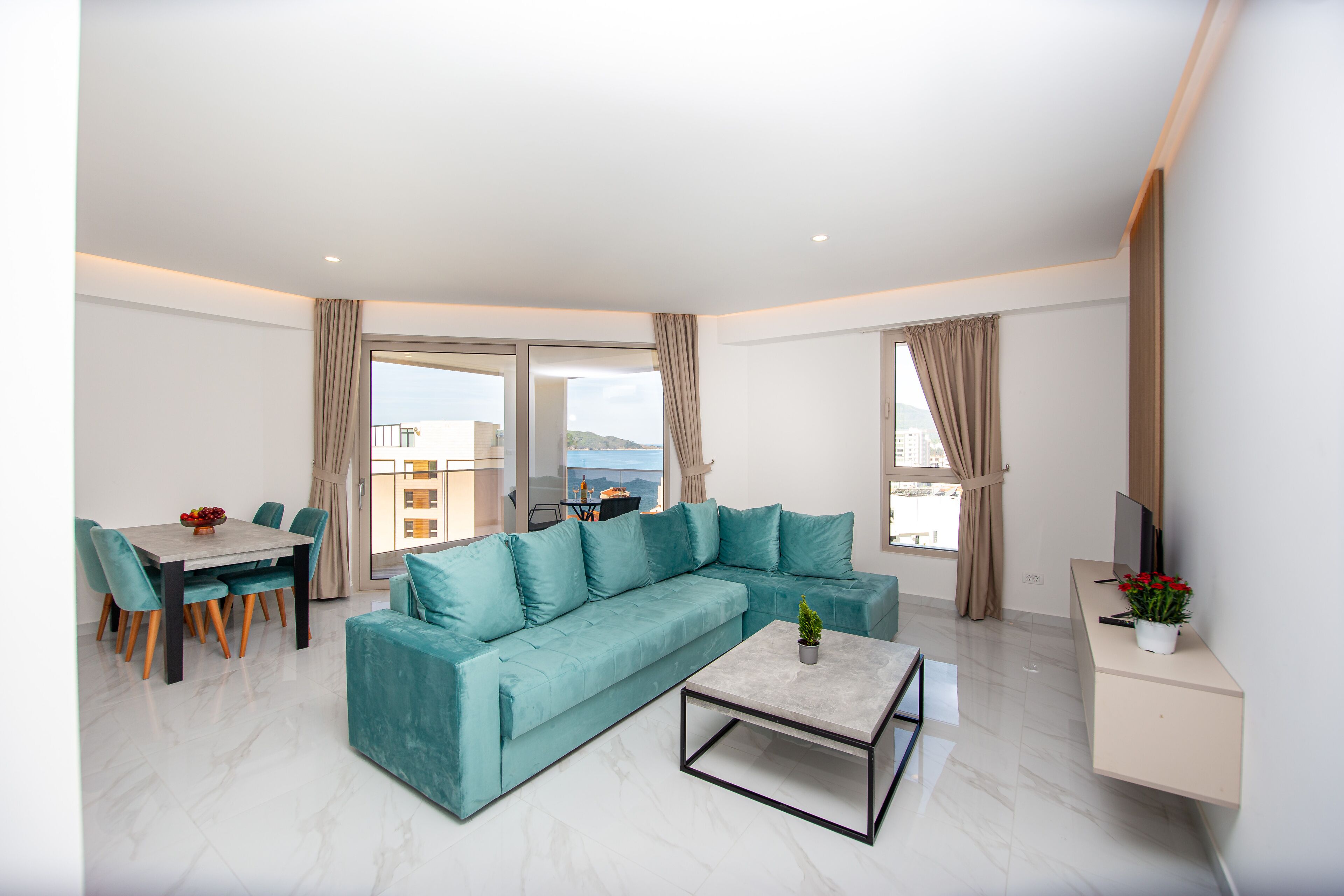 A modern living room with a teal sofa, a dining table with chairs, a coffee table, and a TV on a stand.