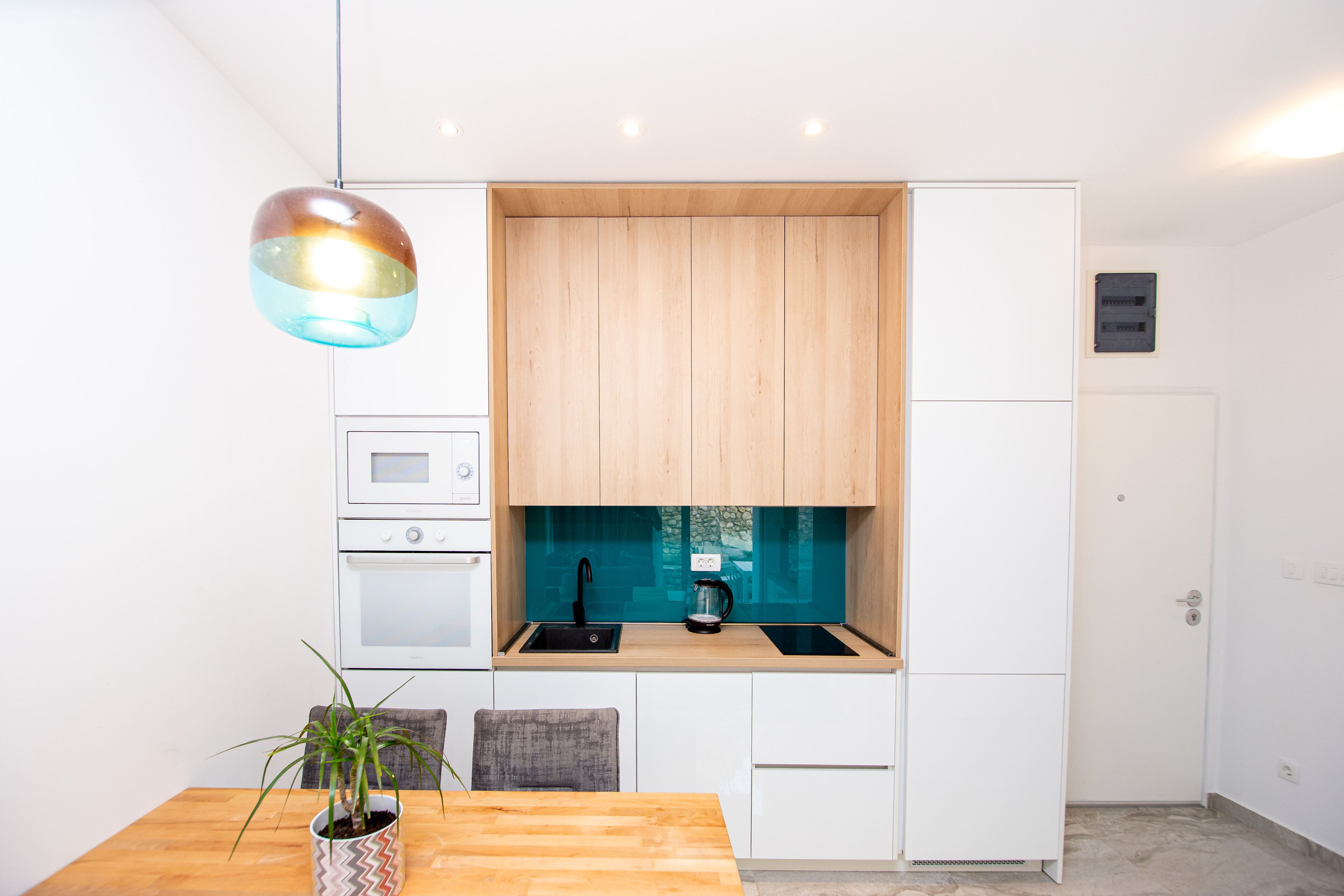 A modern kitchen with white cabinets, a wooden countertop, a sink, and a coffee maker. There is a dining table with chairs and a potted plant.
