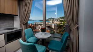 A modern kitchen with a built-in oven, a dining area with two teal chairs and a small table, and a large window offering a city view. - IN Property Apartments (Becici)