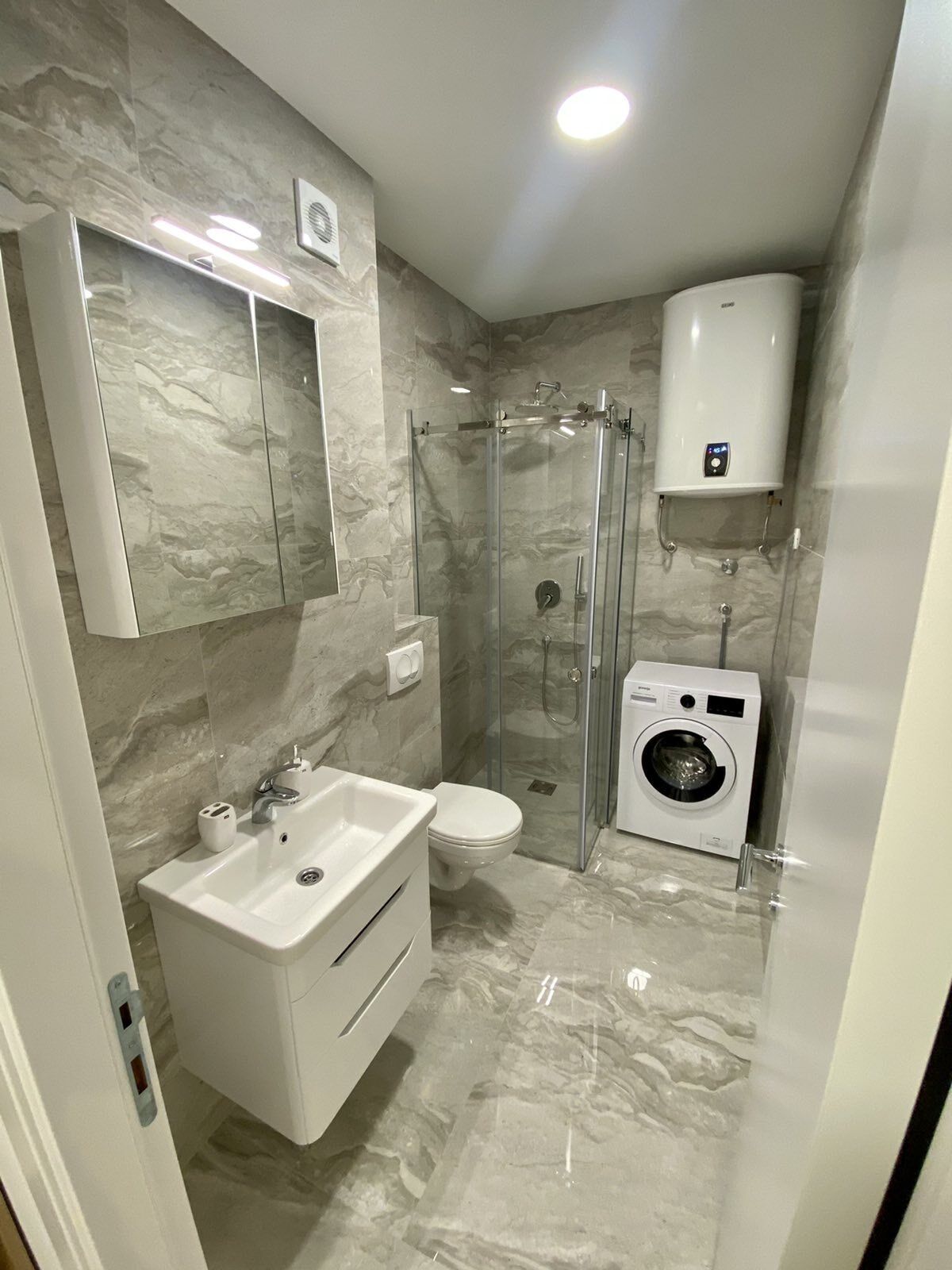 Un baño moderno con ducha de vidrio, lavadora integrada y suelo de mármol.