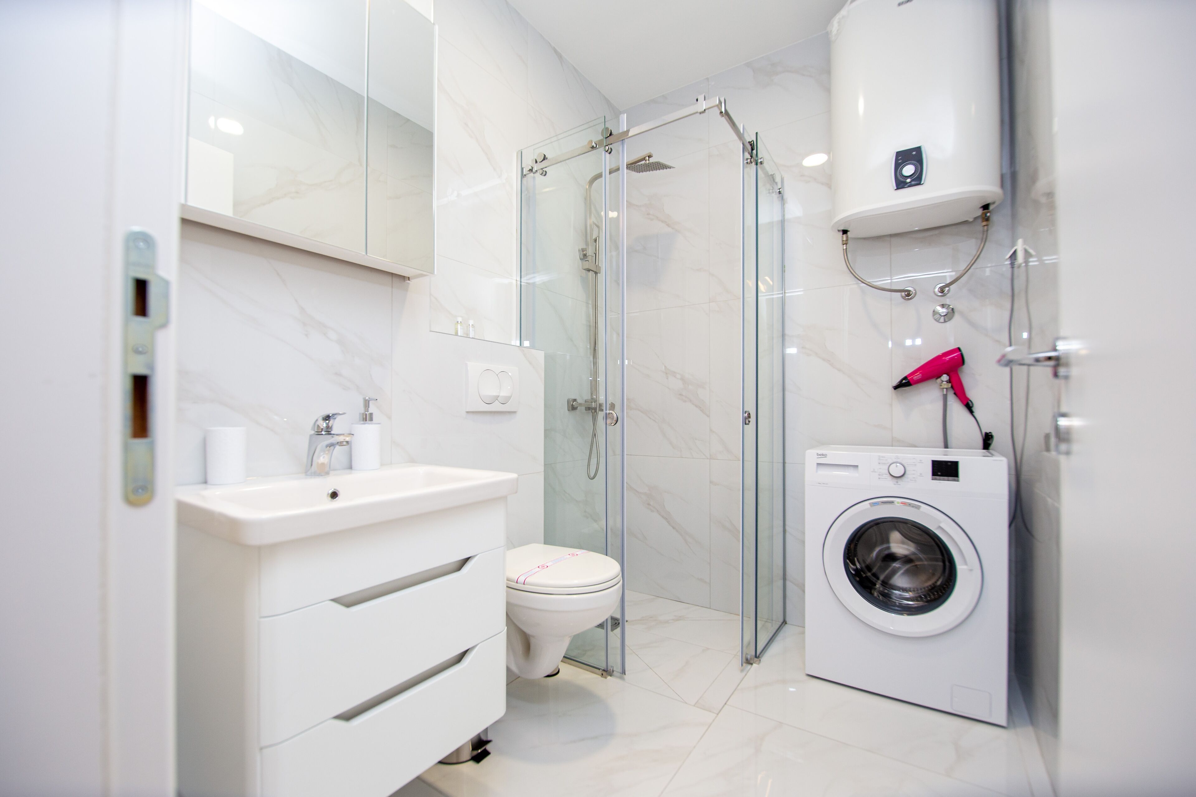 Un baño moderno con ducha envuelta en vidrio, un lavamanos blanco, un inodoro y una lavadora de carga frontal.