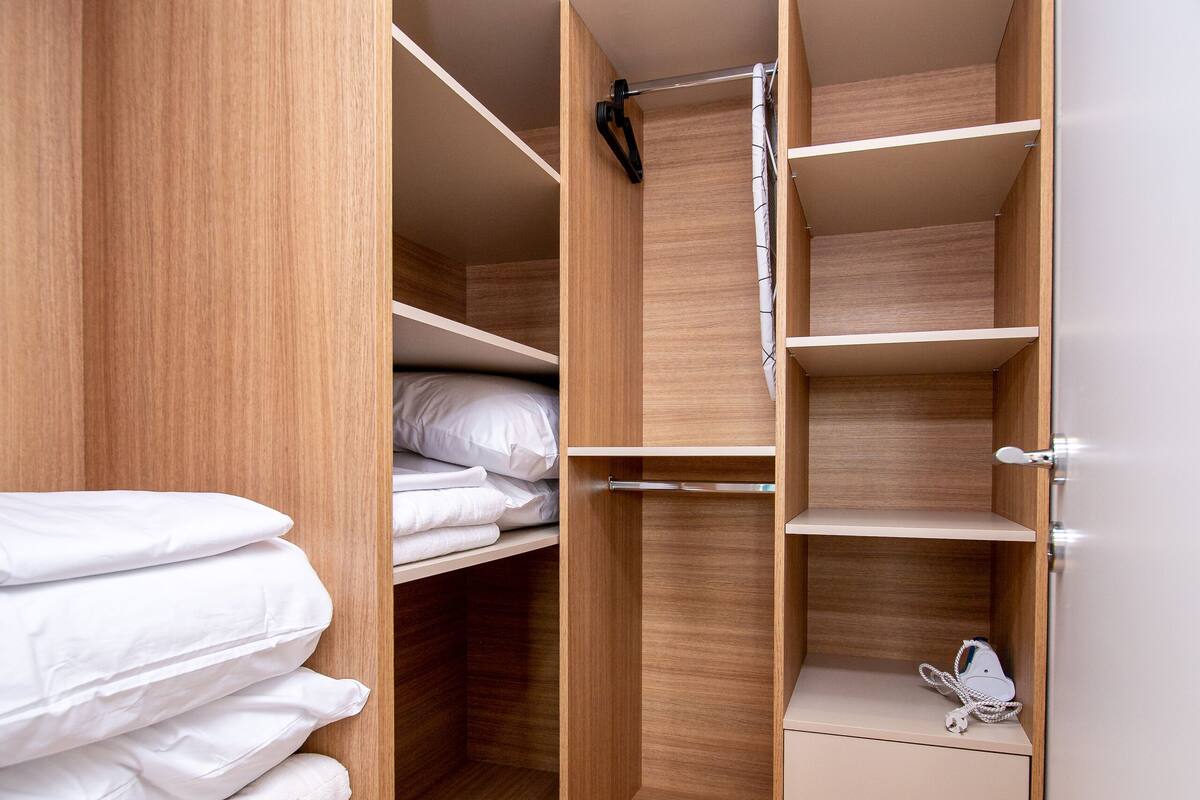 A walk-in closet with wooden shelving and drawers, containing neatly folded white linens and a white electronic device.