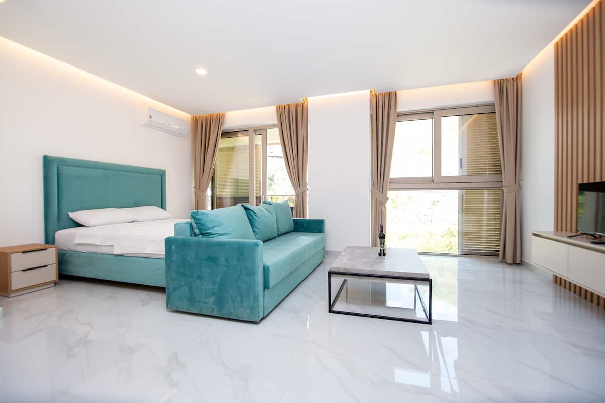 A modern bedroom with a teal sofa, a bed with white linens, a small bedside table, and a TV on a stand.