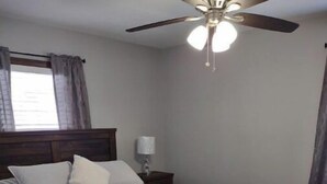 Un dormitorio con cama, mesita de noche, ventilador de techo y ventana con cortinas.