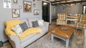 A cozy living room with a sofa, coffee table, and plaid chairs. There are framed pictures on the walls and a wooden ceiling. - Rock Cottage (Penrith)