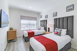 A hotel room with two beds, a window with blinds, a bedside table with a lamp, and framed pictures on the wall.