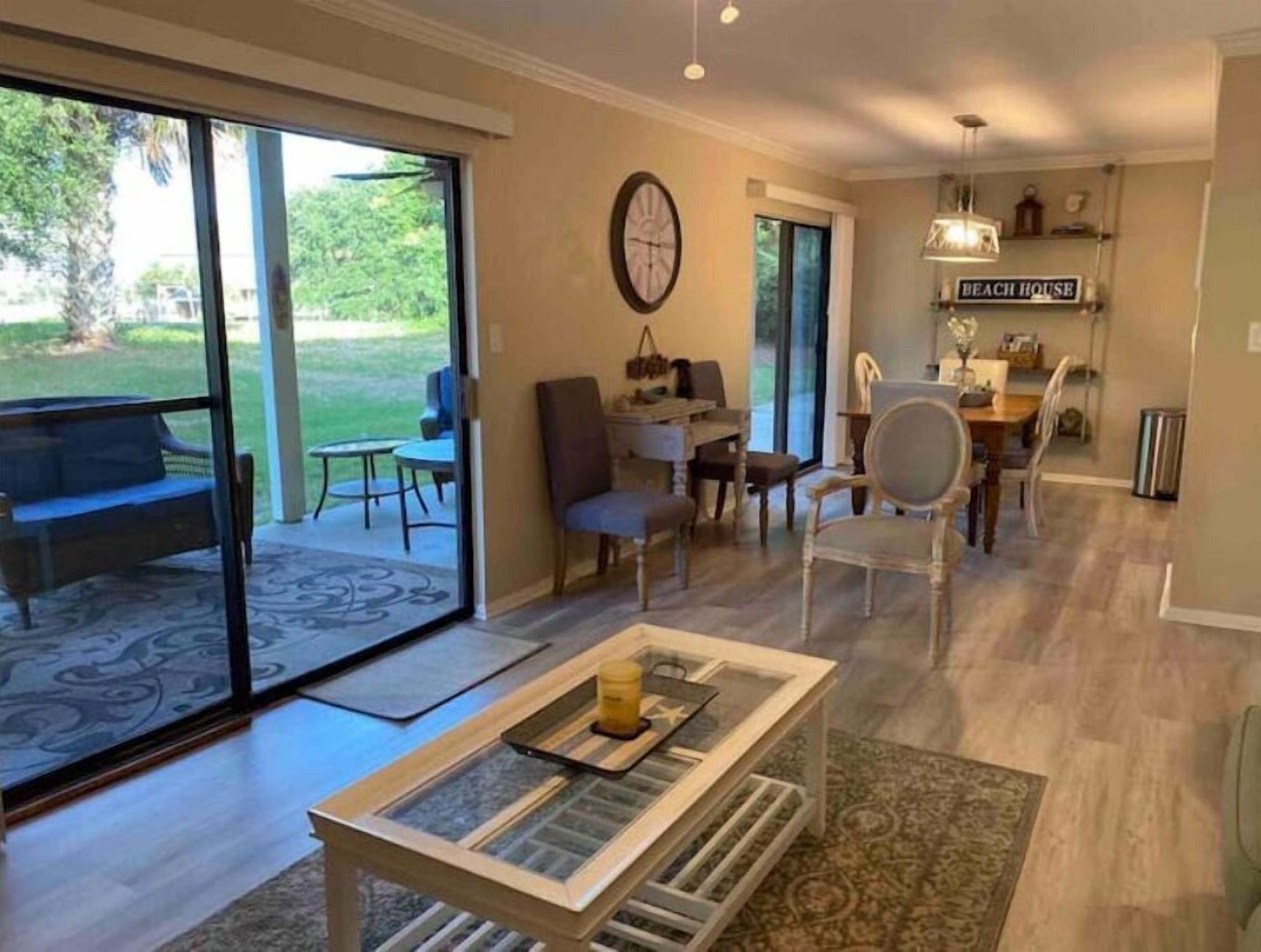 A spacious living area with a large sliding glass door leading to a patio, a dining area with a table and chairs, and a kitchen area with a sign reading "BEACH HOUSE".