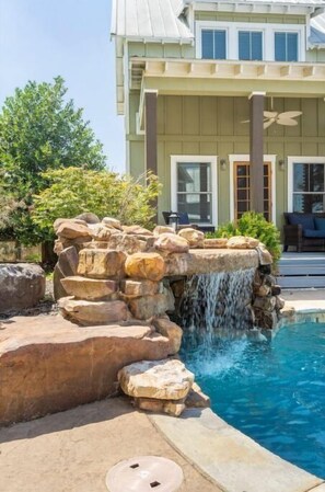 Une piscine agrémentée d’une cascade, entourée de rochers et bordée d’une maison dotée d’une terrasse couverte.