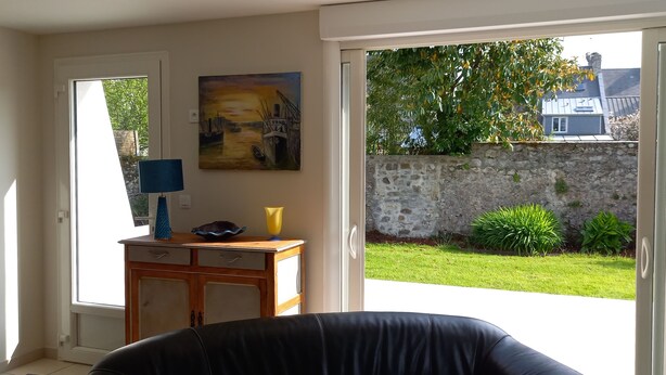A room with a door leading to a garden, a painting on the wall, and a small table with a lamp and vase. - Maison de Campagne Proche mer - 5 Voyageurs (Regnéville-sur-Mer)