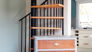 A spiral staircase with wooden steps and black railings, a white dresser with drawers, and a black chair in a room with tiled flooring and a window. - Maison de Campagne Proche mer - 5 Voyageurs (Regnéville-sur-Mer)