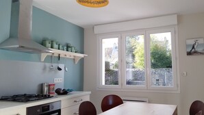 A kitchen with a stove, cabinets, a window, and a dining table with chairs. - Maison de Campagne Proche mer - 5 Voyageurs (Regnéville-sur-Mer)