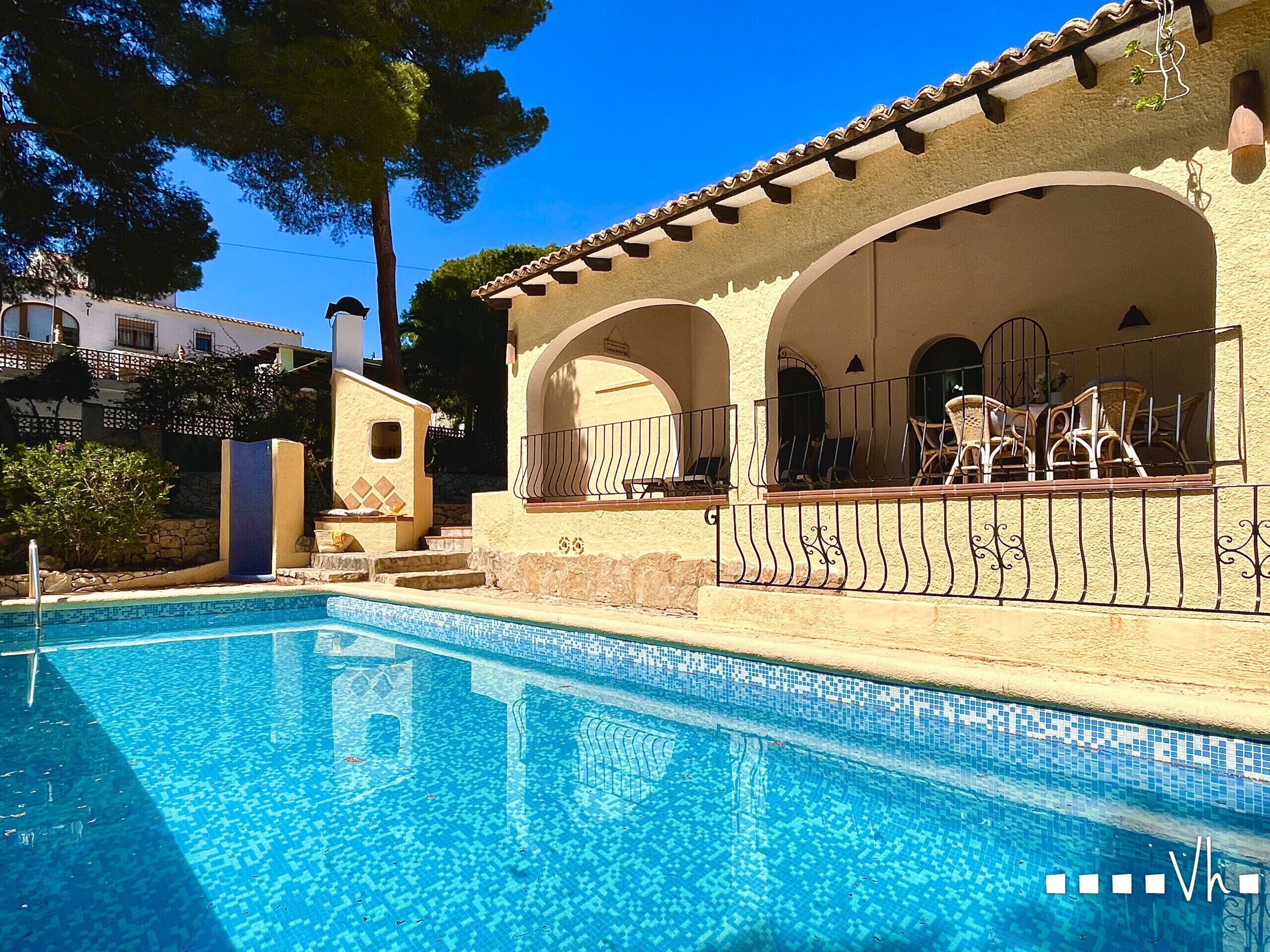 A swimming pool with tiled edges, a covered outdoor seating area with chairs, and a building with arches and a tiled roof.