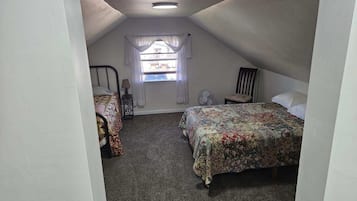 A small, sloped-roofed attic bedroom with two beds, a window with sheer curtains, a standing fan, and a chair.