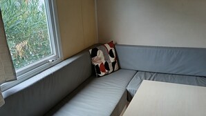 A room with a built-in bench along a window, a table, and a shelf with decorative items. - Bungalow in a comfortable 3-star campsite. Accepted animals. (Argelès-sur-Mer)