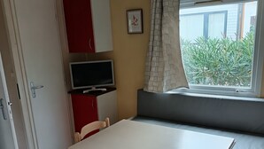 A room with a white table, wooden chairs, a red cabinet, a TV, a window with a view of greenery, and a ceiling with exposed beams. - Bungalow in a comfortable 3-star campsite. Accepted animals. (Argelès-sur-Mer)