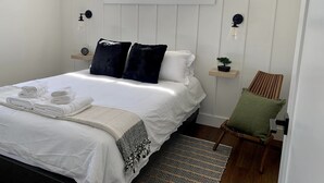 Una cama bien tendida con sábanas blancas y dos almohadas oscuras, una mesita de noche con un reloj, una ventana sobre la cama, una silla con cojín verde y una alfombra estampada en el suelo.