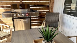 Una mesa de madera con una bandeja que sostiene una planta en maceta, una silla de mimbre y una parrilla al fondo.
