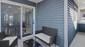 Uma área de varanda coberta com duas cadeiras de vime e uma pequena mesa, uma porta de vidro deslizante que dá para o interior e paredes externas com revestimento horizontal.
