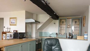 A compact kitchen with a wooden countertop, a stove, a microwave, and a sink. There is a chair and a small table in the foreground. - Gulls Hatch (Maryport)
