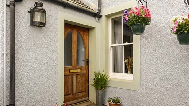 A quaint cottage with a wooden door, a window with a white frame, and hanging flower baskets. - Gulls Hatch (Maryport)