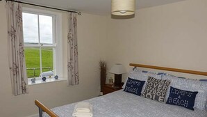 A bedroom with a bed, a window with curtains, a bedside table with a lamp, and a view of the outdoors. - Gulls Hatch (Maryport)